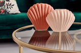 Zwei muschelförmige Vasen in Rosa- und Korallentönen auf einem runden Glastisch mit goldfarbenem Rahmen, im Hintergrund ein grünes Sofa. Perspektive: Nahaufnahme von schräg oben.