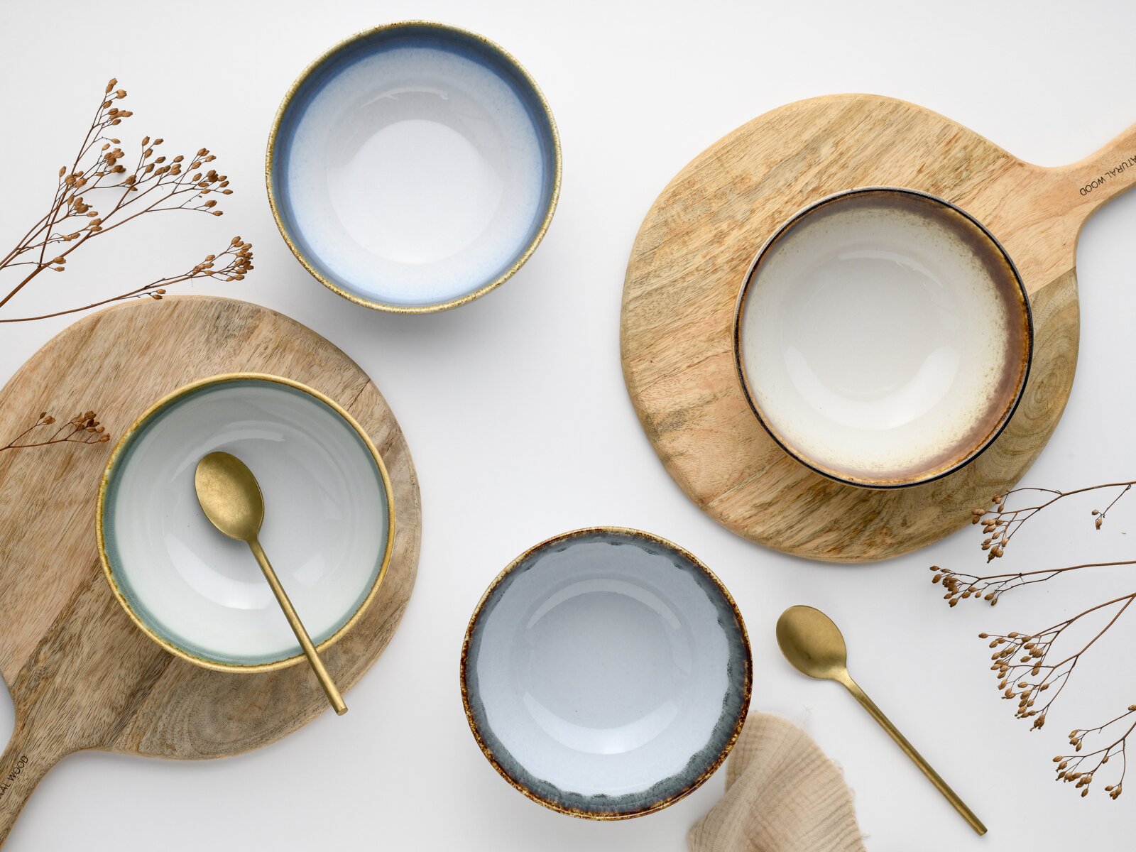 Draufsicht (Flatlay) auf vier glasierte Keramikschalen in Blau- und Beigetönen mit goldbraunem Rand auf Holz-Servierbrettern, dazu zwei goldfarbene Löffel und getrocknete Zweige auf weißem Hintergrund