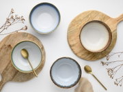 Draufsicht (Flatlay) auf vier glasierte Keramikschalen in Blau- und Beigetönen mit goldbraunem Rand auf Holz-Servierbrettern, dazu zwei goldfarbene Löffel und getrocknete Zweige auf weißem Hintergrund