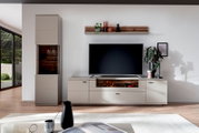Frontale Perspektive: modernes Wohnzimmer-Entertainment-Set mit TV auf niedrigem Sideboard in Taupe mit Holzoptik-Akzenten, links hoher Vitrinenschrank und darüber Wandboard mit Deko vor weißer Wand.