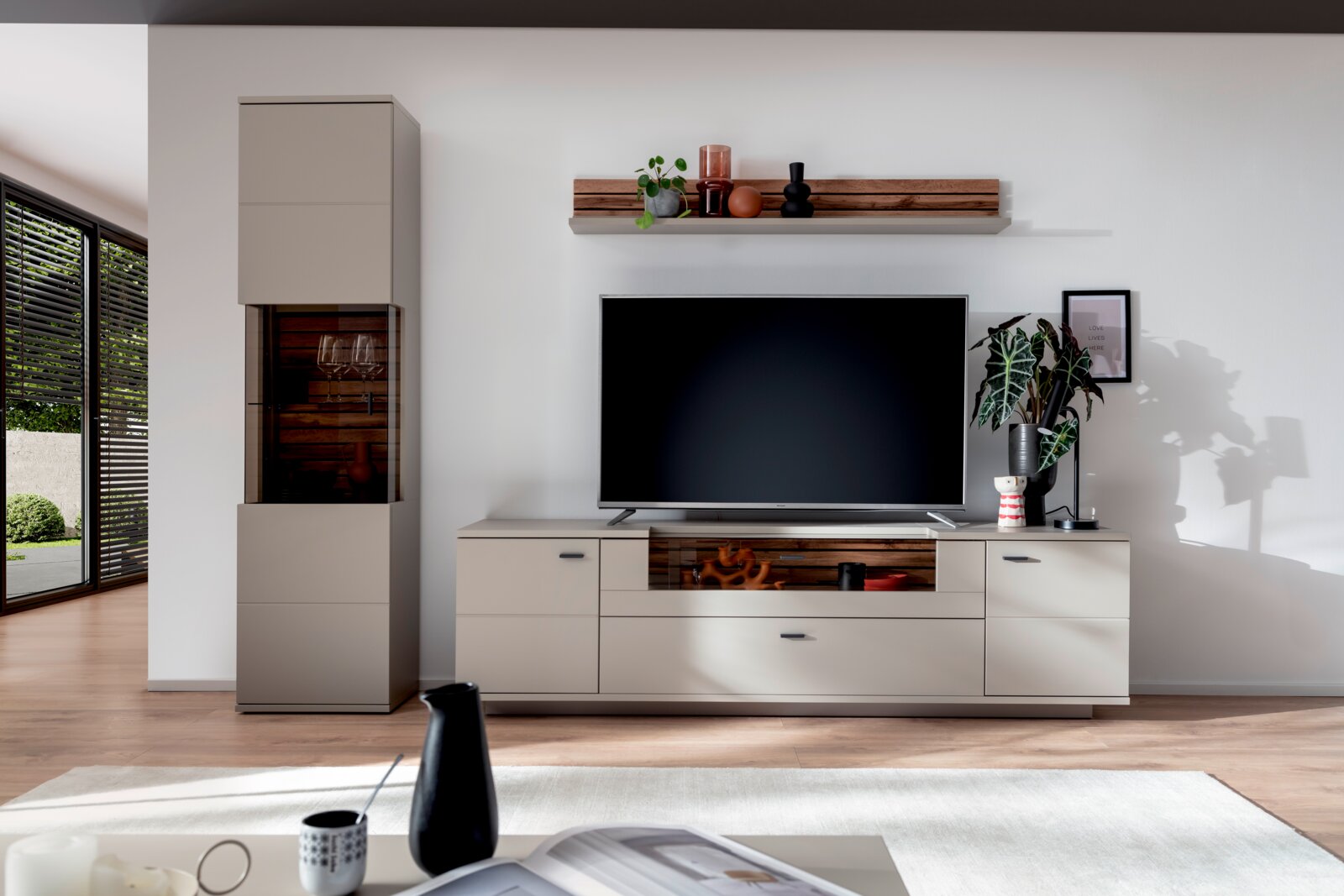 Zurbrüggen Wandcombinatie LÜTTICH Vooraanzicht: moderne entertainmentset voor de woonkamer met tv op een laag dressoir in taupe met accenten in houtlook, links een hoge vitrinekast en daarboven een wandplank met decoratie tegen een witte muur.