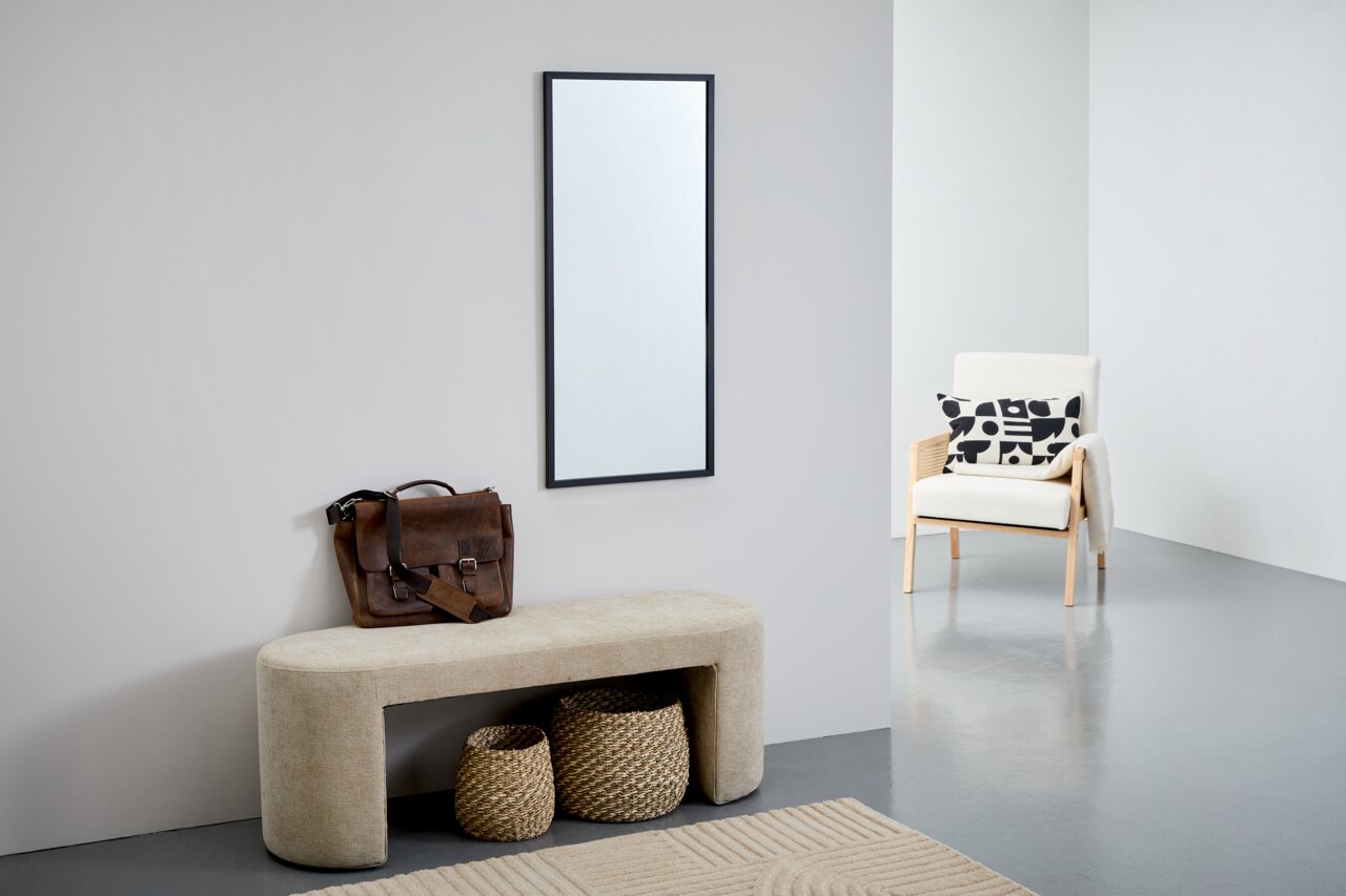 Rechteckiger Spiegel mit schwarzem Rahmen, an einer hellgrauen Wand hängend, im modernen Wohnambiente mit Bank, Korb und Sessel. Perspektive: Frontalansicht.