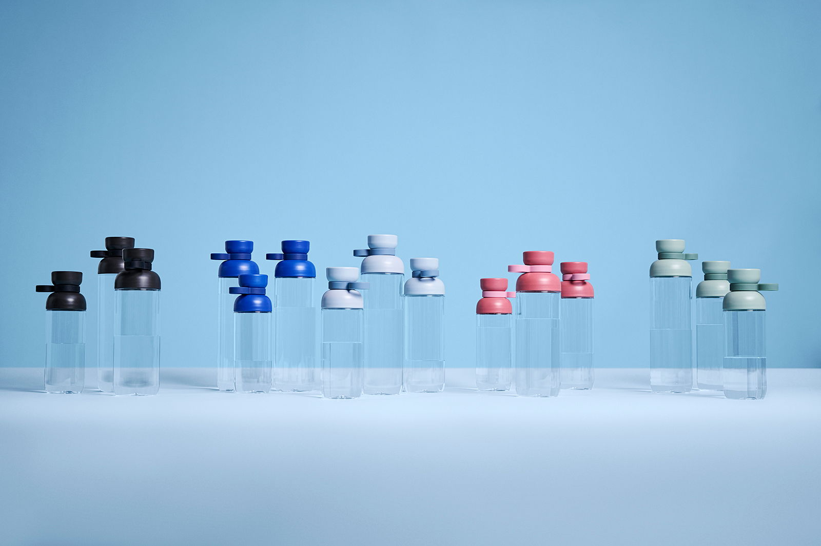 Mehrere Trinkflaschen in verschiedenen Farben, darunter Blau, Schwarz, Weiß, Rosa und Grün, auf einer ebenen Fläche vor einem blauen Hintergrund. Die Flaschen sind aus transparentem Material mit farbigen Deckeln. Perspektive von vorne.
