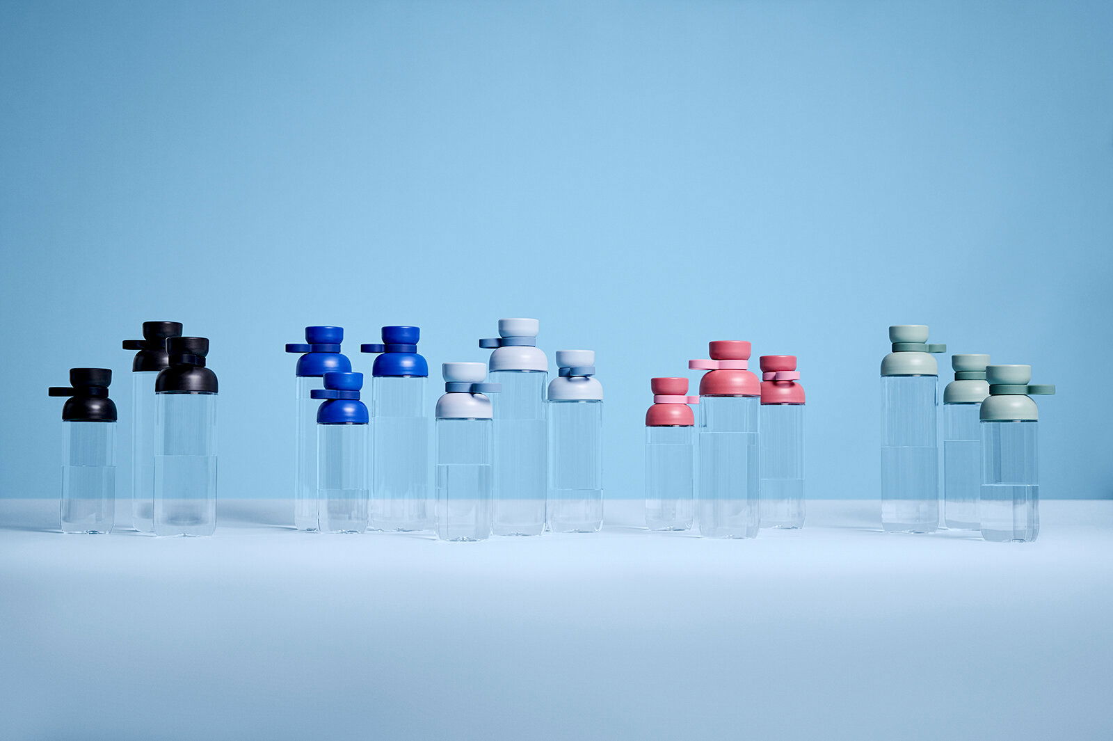Mepal Trinkflasche Nordic Blue VITA Mehrere Trinkflaschen in verschiedenen Farben, darunter Blau, Schwarz, Weiß, Rosa und Grün, auf einer ebenen Fläche vor einem blauen Hintergrund. Die Flaschen sind aus transparentem Material mit farbigen Deckeln. Perspektive von vorne.