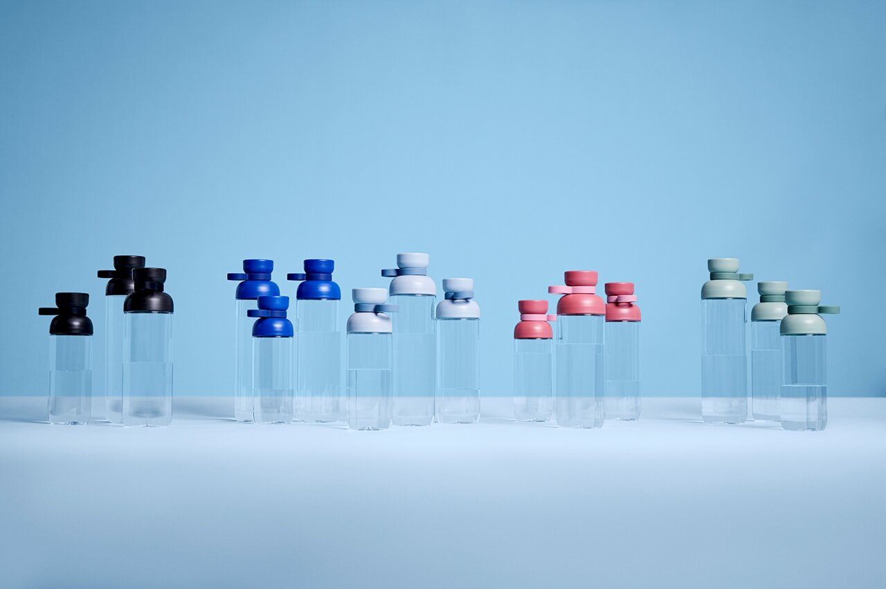 Mehrere Trinkflaschen in verschiedenen Farben, darunter Blau, Schwarz, Weiß, Rosa und Grün, auf einer ebenen Fläche vor einem blauen Hintergrund. Die Flaschen sind aus transparentem Material mit farbigen Deckeln. Perspektive von vorne.