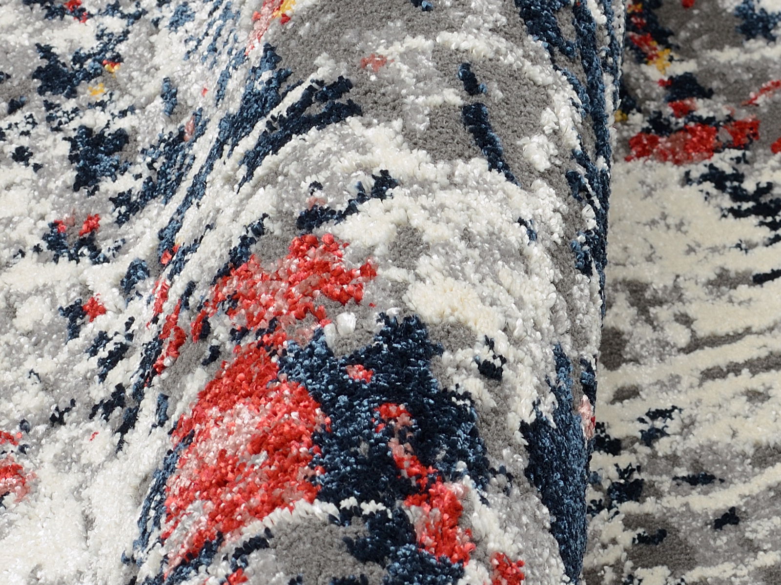 Nahaufnahme eines Designer Teppichs mit abstraktem Muster in Grau, Blau, Rot und Weiß, aus seitlicher Perspektive.