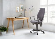 Moderner Schreibtisch aus Holz mit zwei Schubladen, seitliche Perspektive, neben einem ergonomischen Bürostuhl in einem hellen Raum mit großen Fenstern.