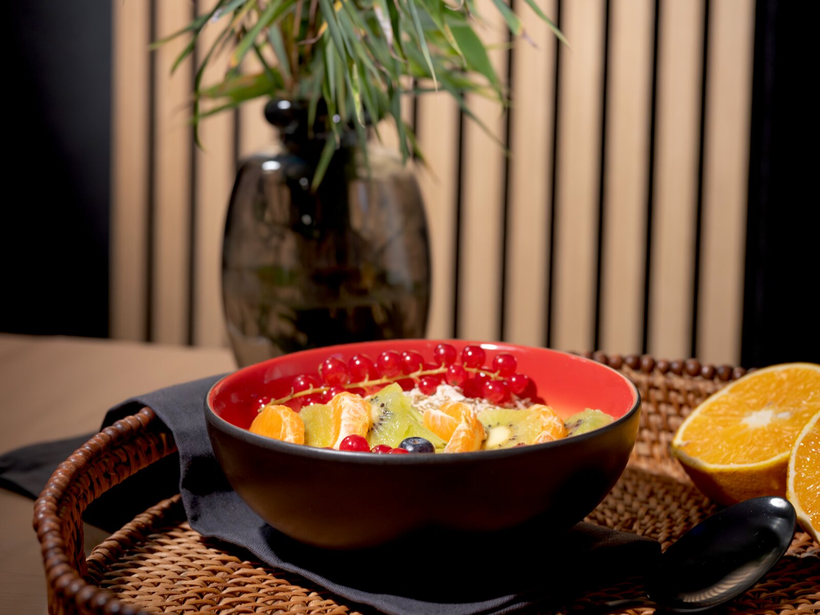 Schwarze Schale mit roter Innenseite, gefüllt mit Müsli und frischen Früchten wie Kiwi, Mandarine, Johannisbeeren und Heidelbeeren, aus leicht erhöhter Frontperspektive auf einem geflochtenen Tablett präsentiert