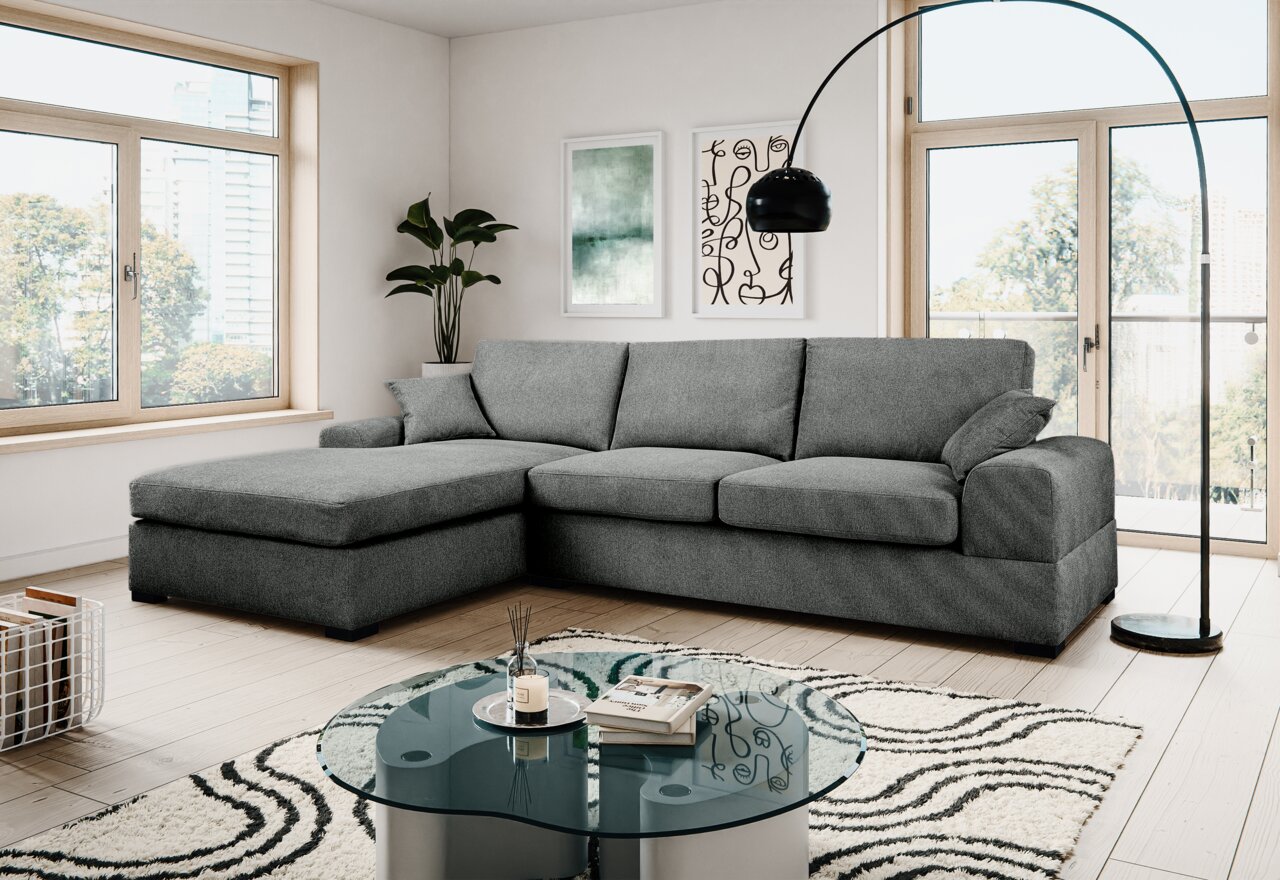 Modernes graues Ecksofa mit linksseitiger Ottomane und drei Sitzkissen in hellem Wohnzimmer, Frontansicht leicht schräg von vorn.