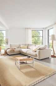 Helles Wohnzimmer mit großer, beiger Rundecke im Vordergrund, von der Seite betrachtet. Ein kleiner Couchtisch steht auf einem Teppich vor der Couch.