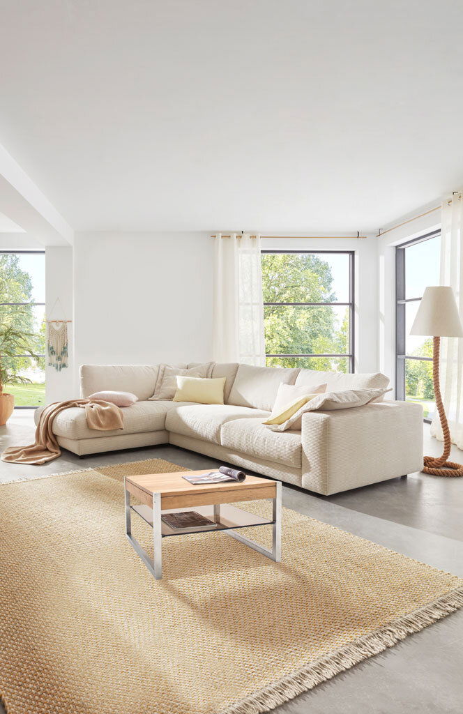 Helles Wohnzimmer mit großer, beiger Rundecke im Vordergrund, von der Seite betrachtet. Ein kleiner Couchtisch steht auf einem Teppich vor der Couch.