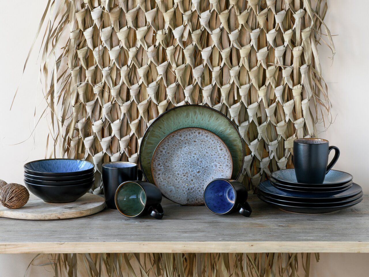 Kombiservice Bali 16-tlg. in einer stilvollen Präsentation, bestehend aus Tellern, Schalen und Tassen in verschiedenen Blau- und Grüntönen, auf einem Holztisch vor einem geflochtenen Hintergrund. Frontale Perspektive.