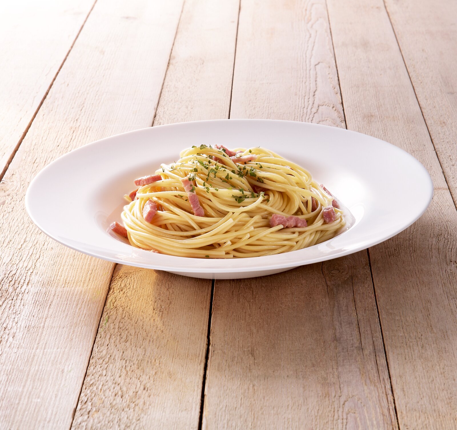 Weiße Pastaschale mit Spaghetti und Speckstreifen, mit Kräutern garniert, auf hellem Holztisch, fotografiert aus leicht erhöhter Schrägansicht von vorne