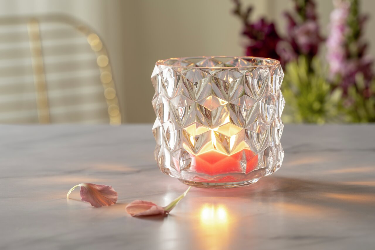 Glas-Tischlicht mit großen Rauten, beleuchtet von einer Kerze, auf einem Tisch mit Blumen im Hintergrund, seitliche Perspektive.