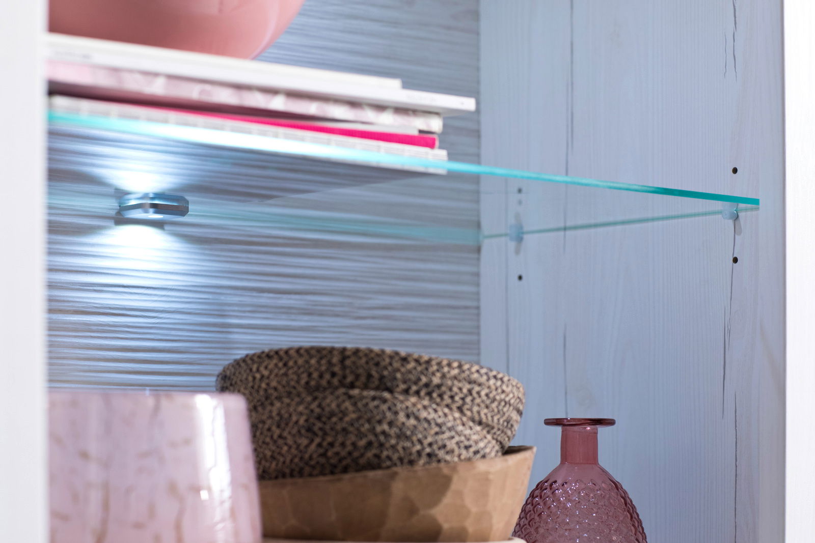 Nahaufnahme eines Regals mit Glasbodenbeleuchtung, dekoriert mit rosa Glasflasche und geflochtenen Körben, seitliche Perspektive.