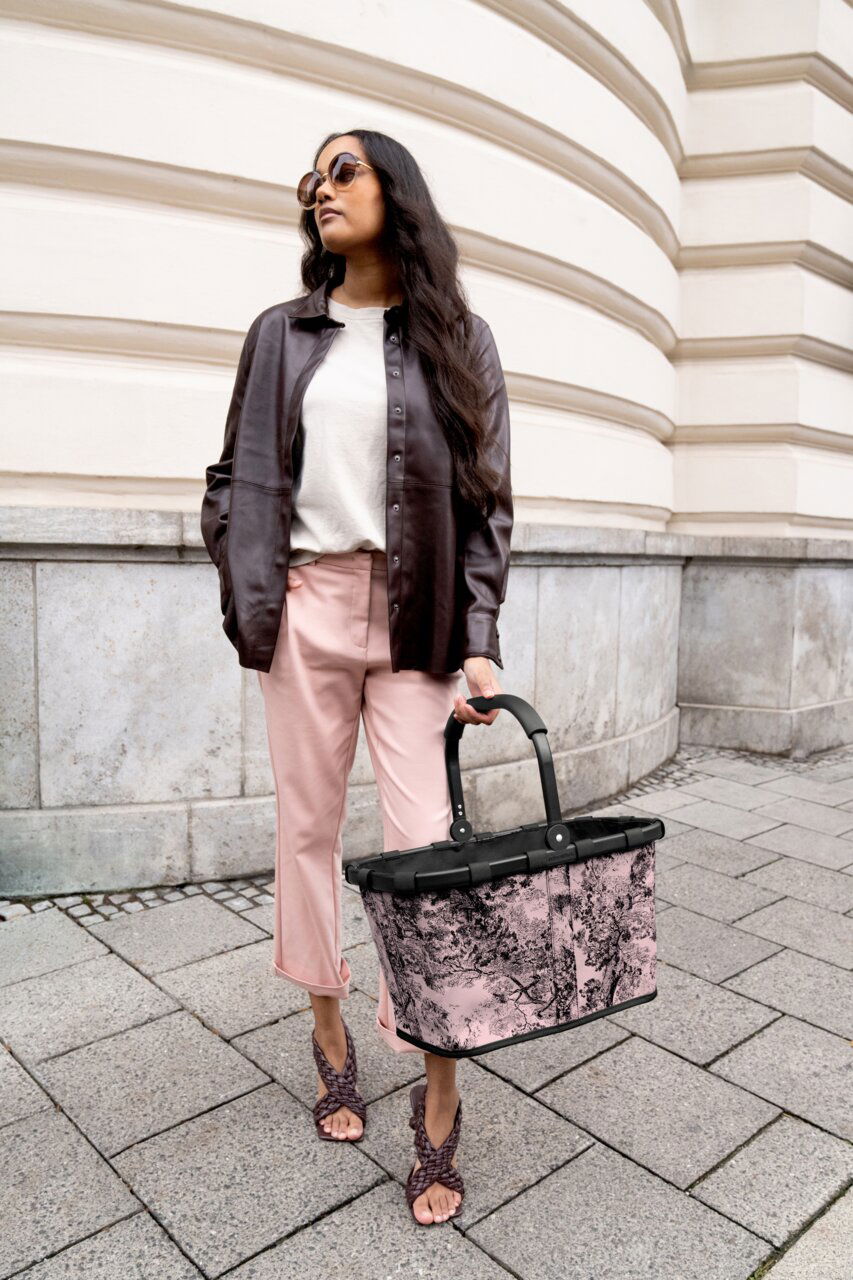 Frau in eleganter Kleidung hält einen stilvollen, rosa gemusterten Einkaufskorb in der Hand, seitliche Perspektive.