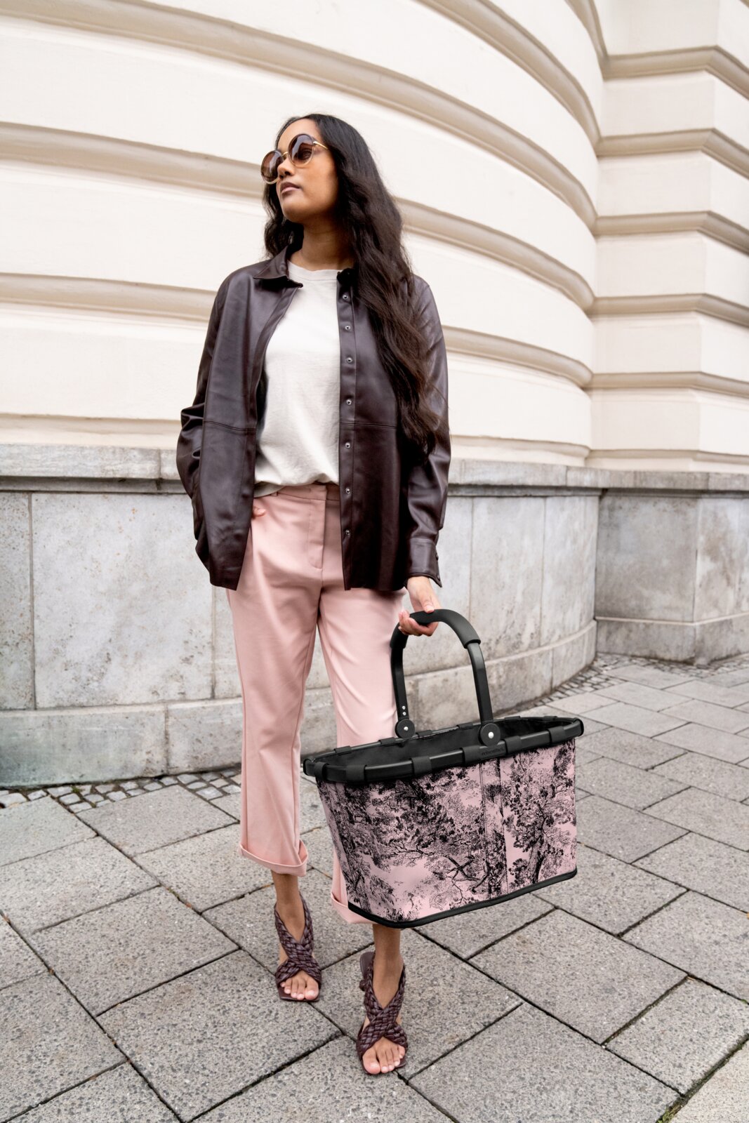 Frau in eleganter Kleidung hält einen stilvollen, rosa gemusterten Einkaufskorb in der Hand, seitliche Perspektive.