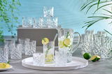 Ein elegantes 18-teiliges Becherset Noblesse mit Kristallgläsern und einem Krug, präsentiert auf einem Tisch mit frischen Limetten und einem modernen, hellen Hintergrund. Perspektive von vorne.
