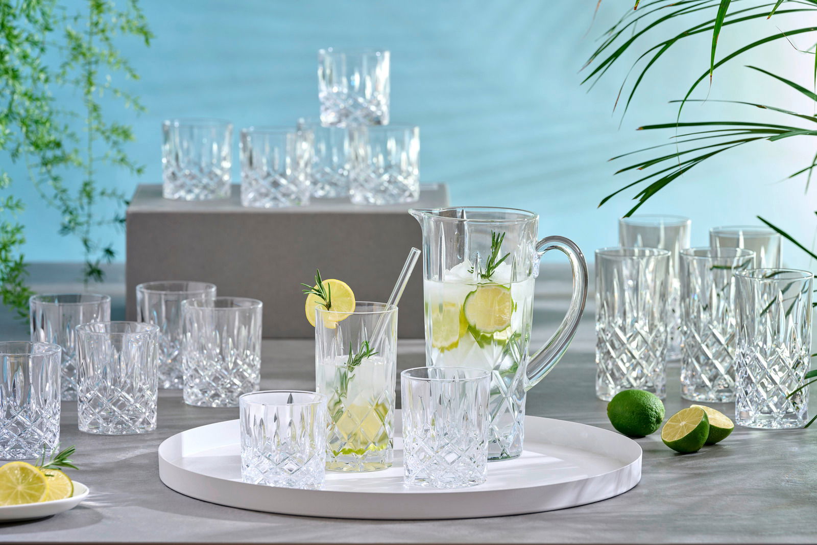 Ein elegantes 18-teiliges Becherset Noblesse mit Kristallgläsern und einem Krug, präsentiert auf einem Tisch mit frischen Limetten und einem modernen, hellen Hintergrund. Perspektive von vorne.