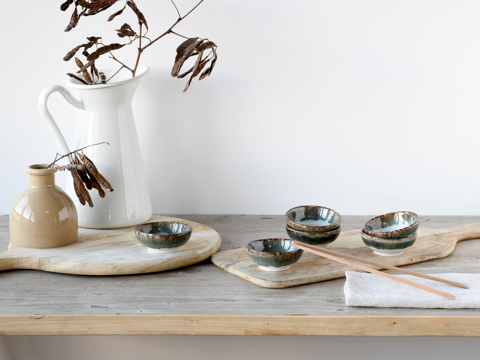 Rustikales Stillleben in leichter Seitenansicht: drei glasierte Keramikschalen in Grün-Blau auf Holzbrett mit Essstäbchen, daneben weiße Kanne und kleine beige Vase mit getrockneten Zweigen auf Holztisch vor hellem Hintergrund.
