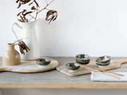 Rustikales Stillleben in leichter Seitenansicht: drei glasierte Keramikschalen in Grün-Blau auf Holzbrett mit Essstäbchen, daneben weiße Kanne und kleine beige Vase mit getrockneten Zweigen auf Holztisch vor hellem Hintergrund.