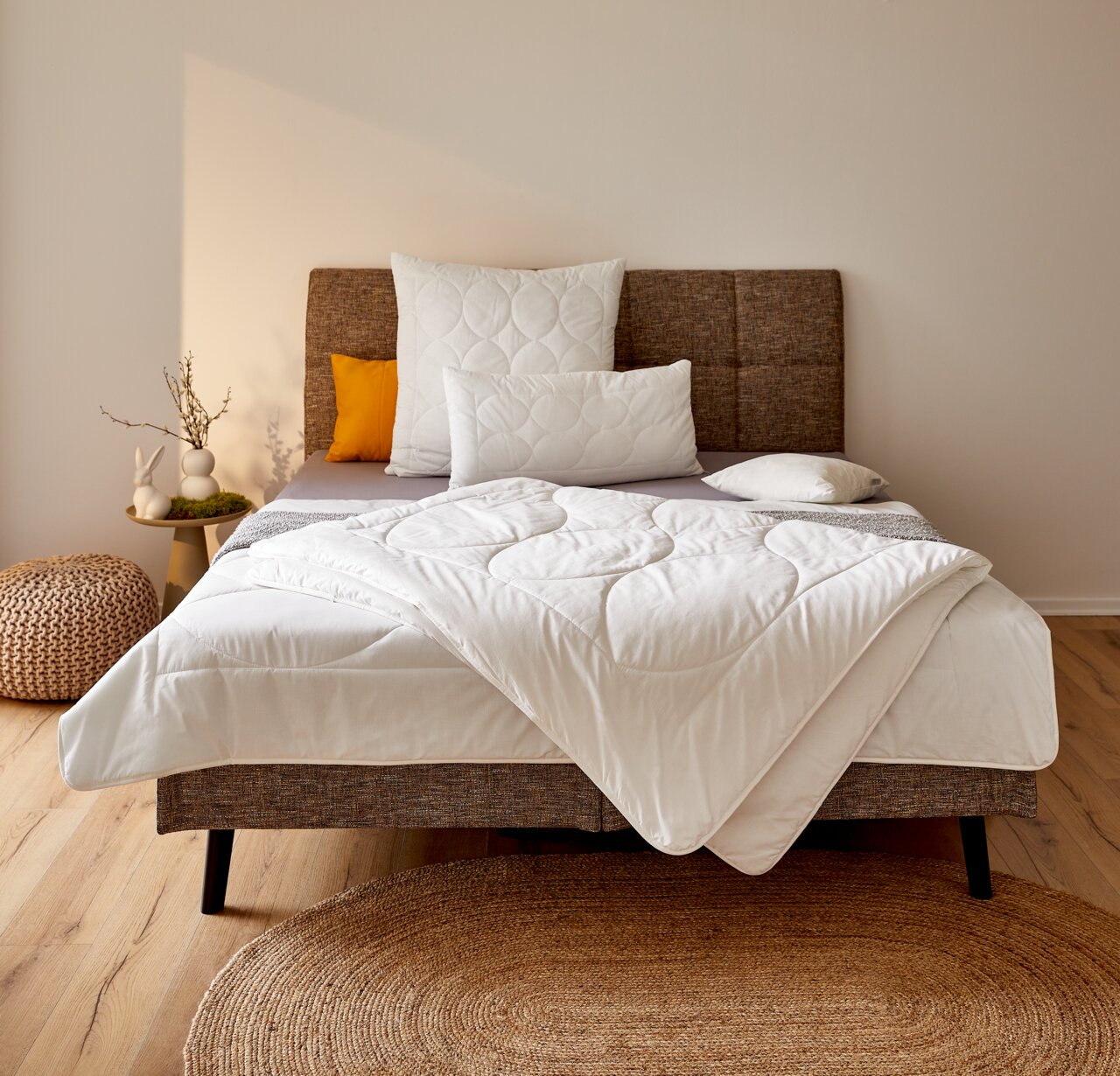 Gemütliches Schlafzimmer mit einem stilvollen Bett, das mit einer weißen Ganzjahresbettdecke in 155x220 cm Größe ausgestattet ist. Die Bettdecke ist elegant auf dem Bett drapiert, umgeben von dekorativen Kissen und einem modernen Kopfteil. Die Perspektive