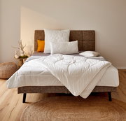 Gemütliches Schlafzimmer mit einem stilvollen Bett, das mit einer weißen Ganzjahresbettdecke in 155x220 cm Größe ausgestattet ist. Die Bettdecke ist elegant auf dem Bett drapiert, umgeben von dekorativen Kissen und einem modernen Kopfteil. Die Perspektive