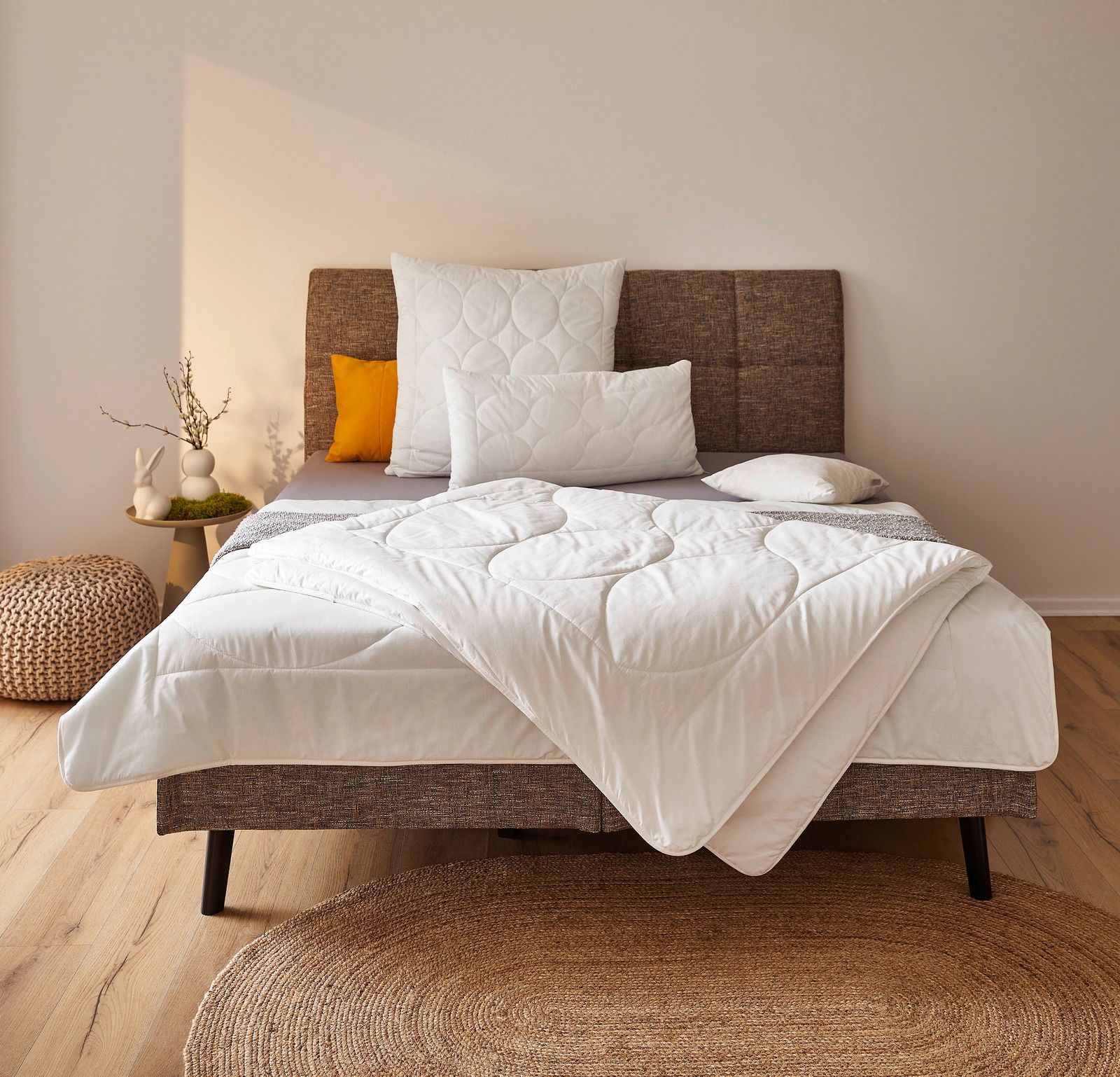 Gemütliches Schlafzimmer mit einem stilvollen Bett, das mit einer weißen Ganzjahresbettdecke in 155x220 cm Größe ausgestattet ist. Die Bettdecke ist elegant auf dem Bett drapiert, umgeben von dekorativen Kissen und einem modernen Kopfteil. Die Perspektive