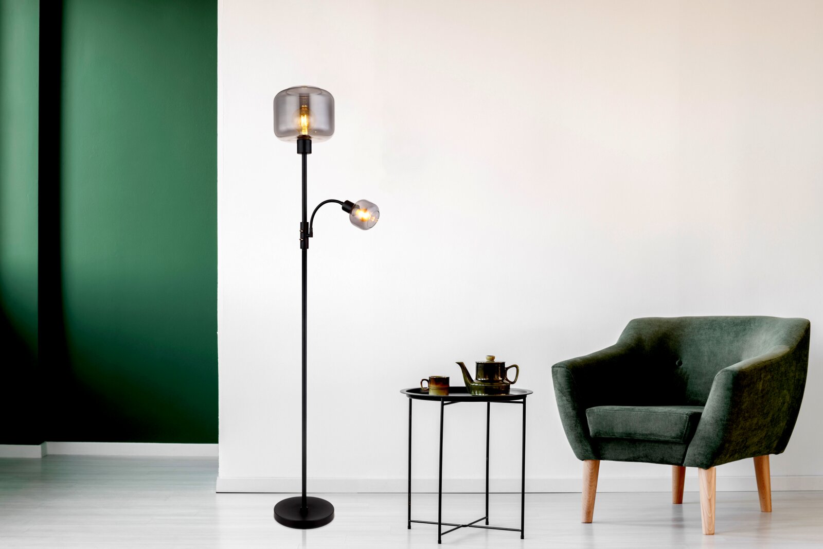 Moderne Stehleuchte mit zwei Leuchtkörpern, einer großen oberen und einer kleineren seitlichen, in einem stilvollen Wohnzimmer mit grünem Sessel und Beistelltisch. Perspektive von vorne.