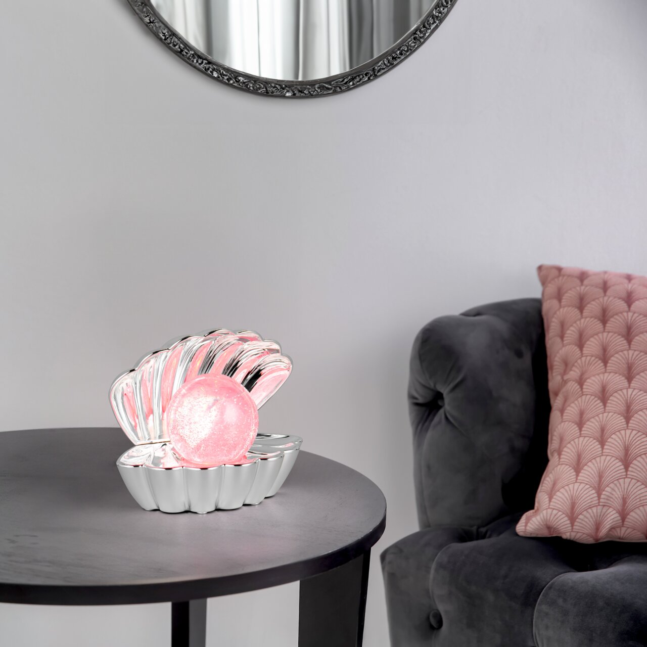 Reality Leuchten LED-Tischleuchte SHELLY Tischleuchte in Form einer geöffneten Muschel mit einer leuchtenden rosa Kugel, auf einem Tisch neben einem grauen Sessel mit rosa Kissen, seitliche Perspektive.