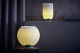 Zwei kugelförmige Lampen mit gebürstetem Silberfuß, die warmes gelbes Licht ausstrahlen. Eine Lampe steht im Vordergrund auf einem Tisch, die andere im Hintergrund auf einem Regal.