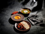 Schräg von oben fotografiertes Food-Arrangement auf dunkler Steinoberfläche: drei braune Keramikteller mit cremiger Suppe, Salat mit Burrata und Kirschtomaten sowie Fischfilet mit heller Sauce und Gemüsebeilage, daneben Tomatenrispe und graues Tuch.