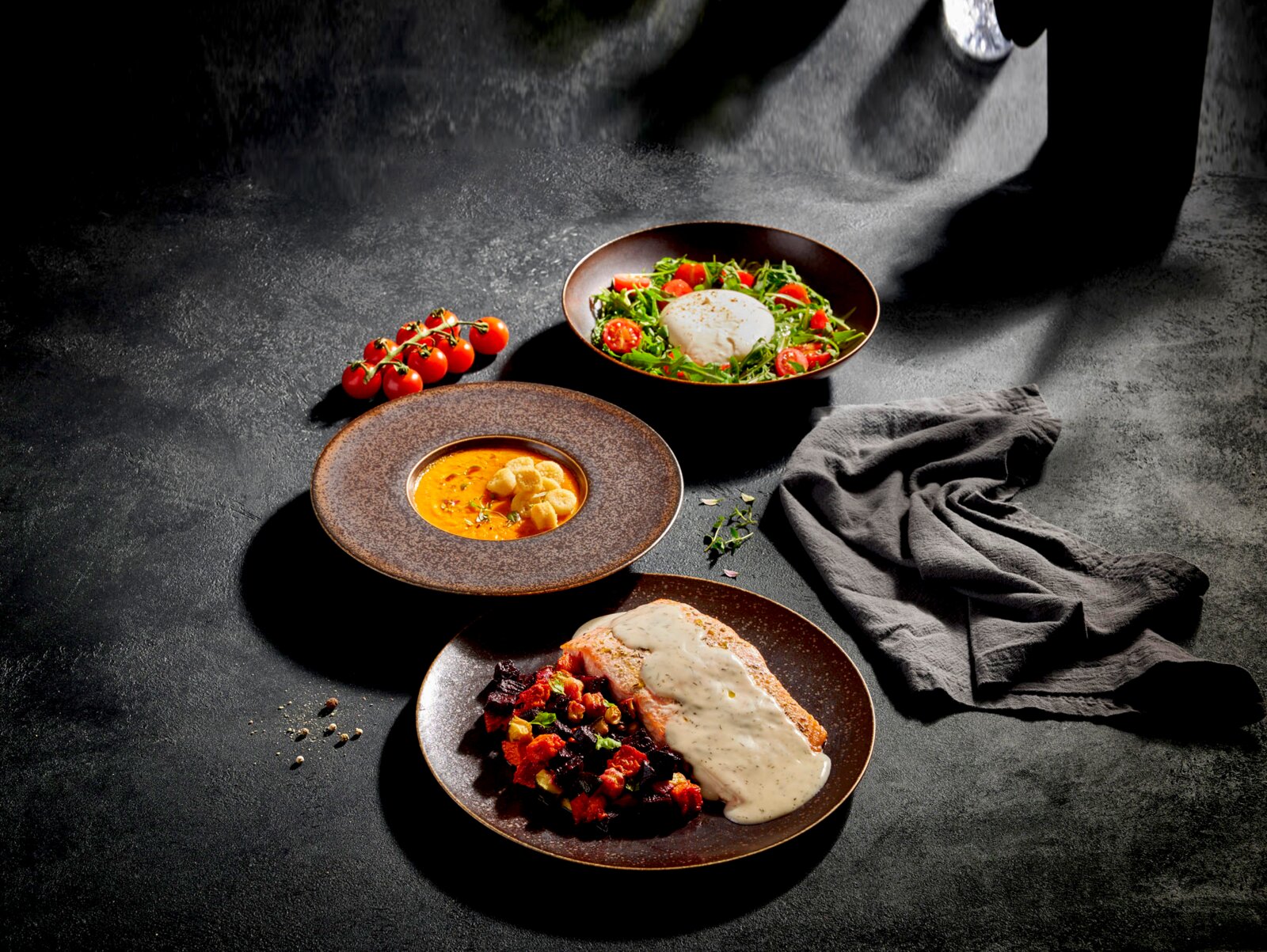 Schräg von oben fotografiertes Food-Arrangement auf dunkler Steinoberfläche: drei braune Keramikteller mit cremiger Suppe, Salat mit Burrata und Kirschtomaten sowie Fischfilet mit heller Sauce und Gemüsebeilage, daneben Tomatenrispe und graues Tuch.