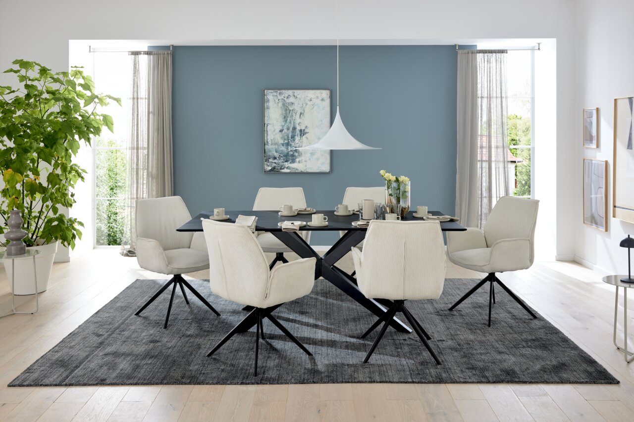 Musterring Eettafel MR TIAMA Eetkamer met een moderne eettafel en zes gestoffeerde stoelen met armleuningen in een lichte ruimte met grote ramen, gezien vanaf de voorkant.