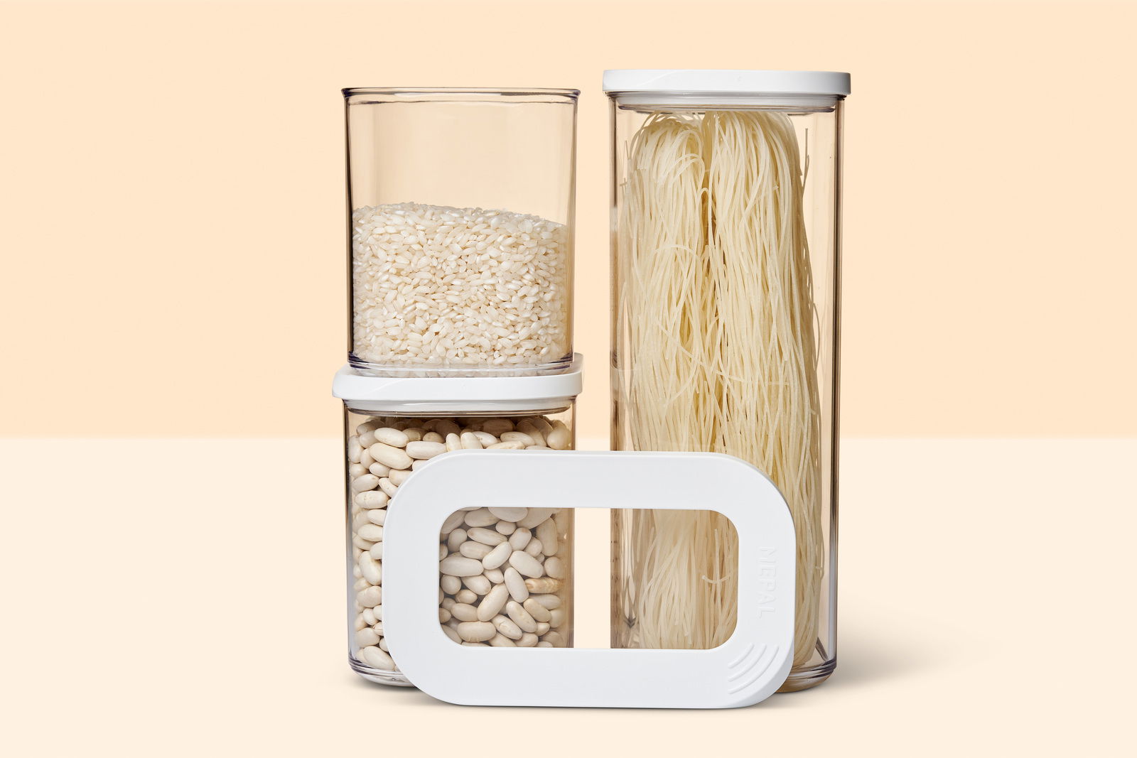 Drei transparente Vorratsbehälter mit weißen Deckeln, gefüllt mit Reis, Bohnen und Nudeln, aus der Frontalperspektive.