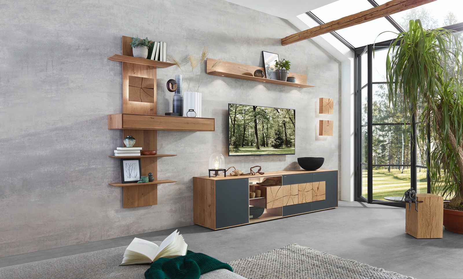 Moderne Wandkombination Valnatura LAGO in einem hellen Wohnzimmer mit großen Fenstern, bestehend aus hölzernen Regalen und Schränken mit dekorativen Elementen. Perspektive von schräg vorne.