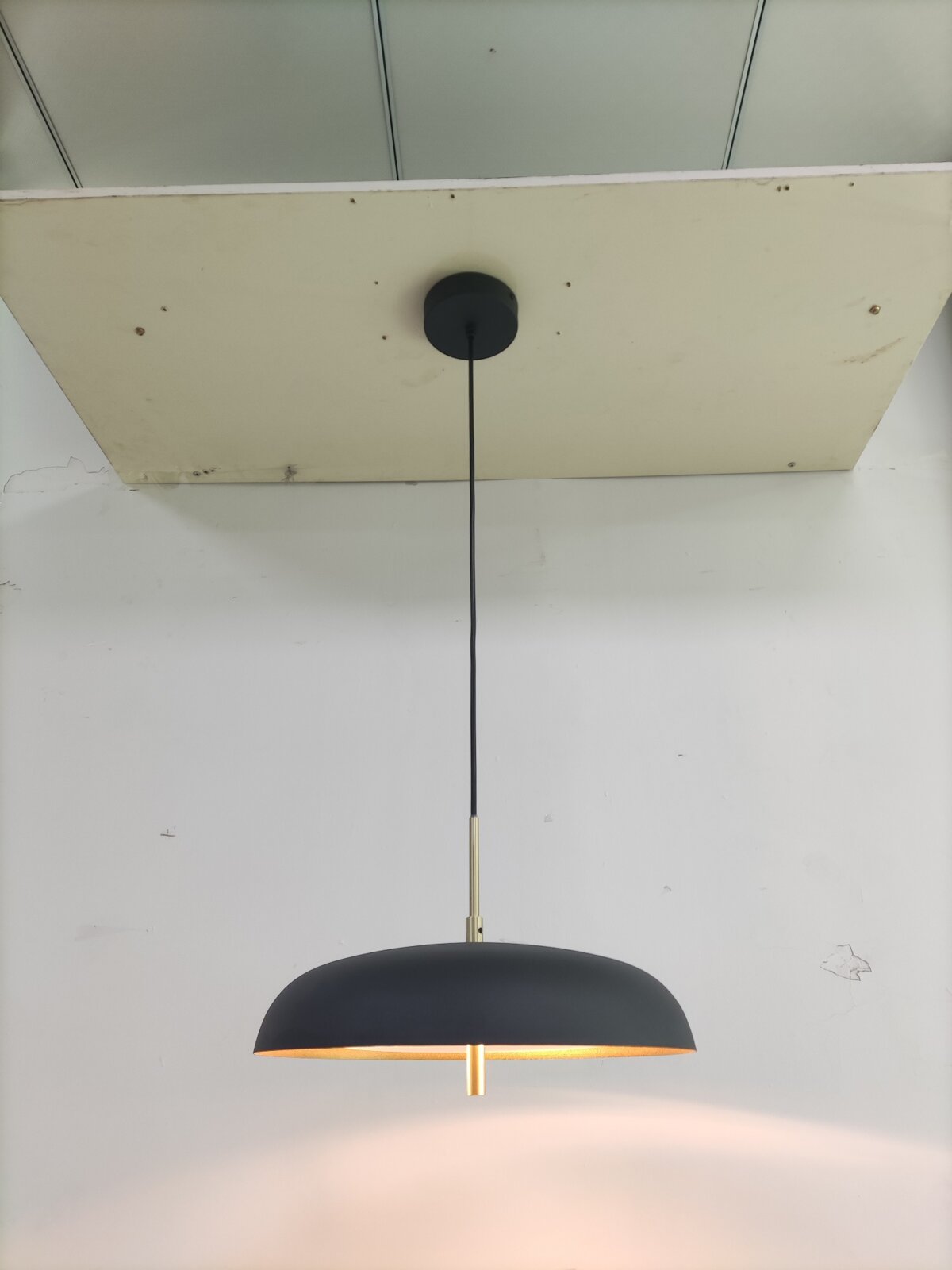 Novel HÄNGELEUCHTE Moderne Pendelleuchte mit flachem, mattschwarzem Schirm und Messing-Details, warm leuchtend, frontal von unten fotografiert.