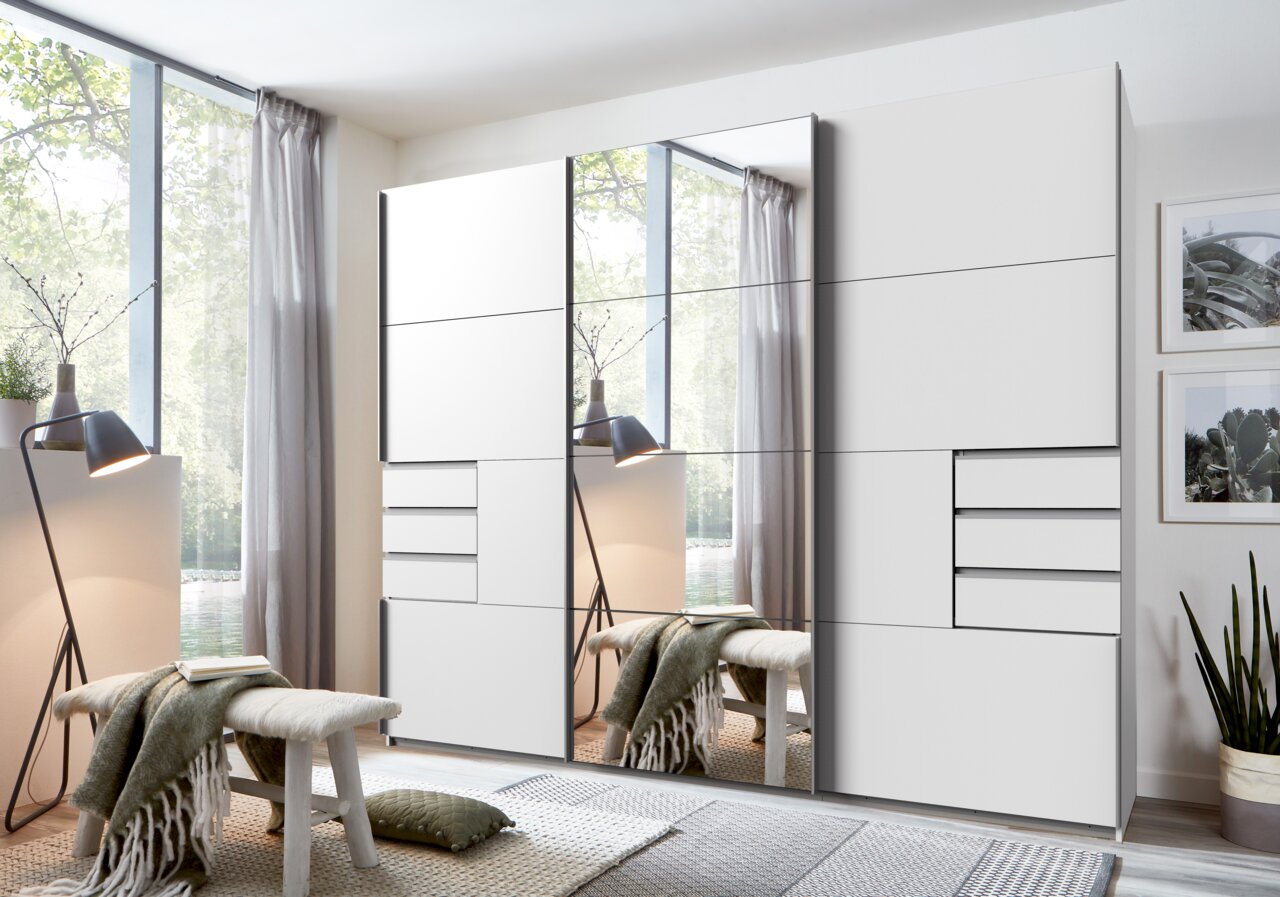 Moderner Schwebetürenschrank mit drei Türen, teilweise mit Spiegeln, in einem hellen Raum mit Blick auf ein großes Fenster.