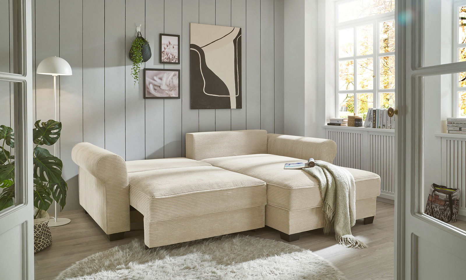 Beige Ecksofa in einem hellen Wohnzimmer mit großen Fenstern, dekoriert mit Pflanzen und moderner Kunst, aus der Perspektive von vorne links.