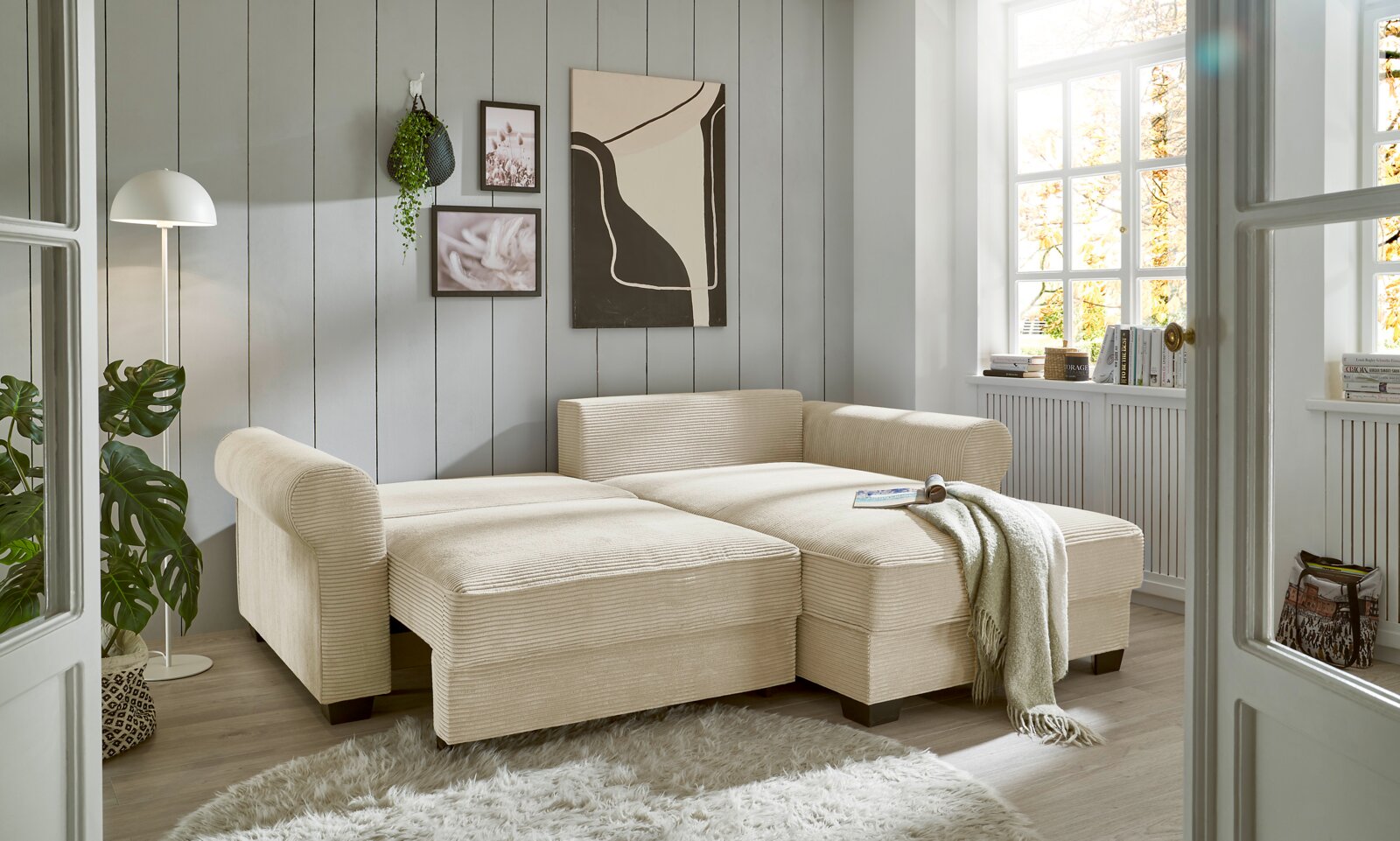 Beige Ecksofa in einem hellen Wohnzimmer mit großen Fenstern, dekoriert mit Pflanzen und moderner Kunst, aus der Perspektive von vorne links.