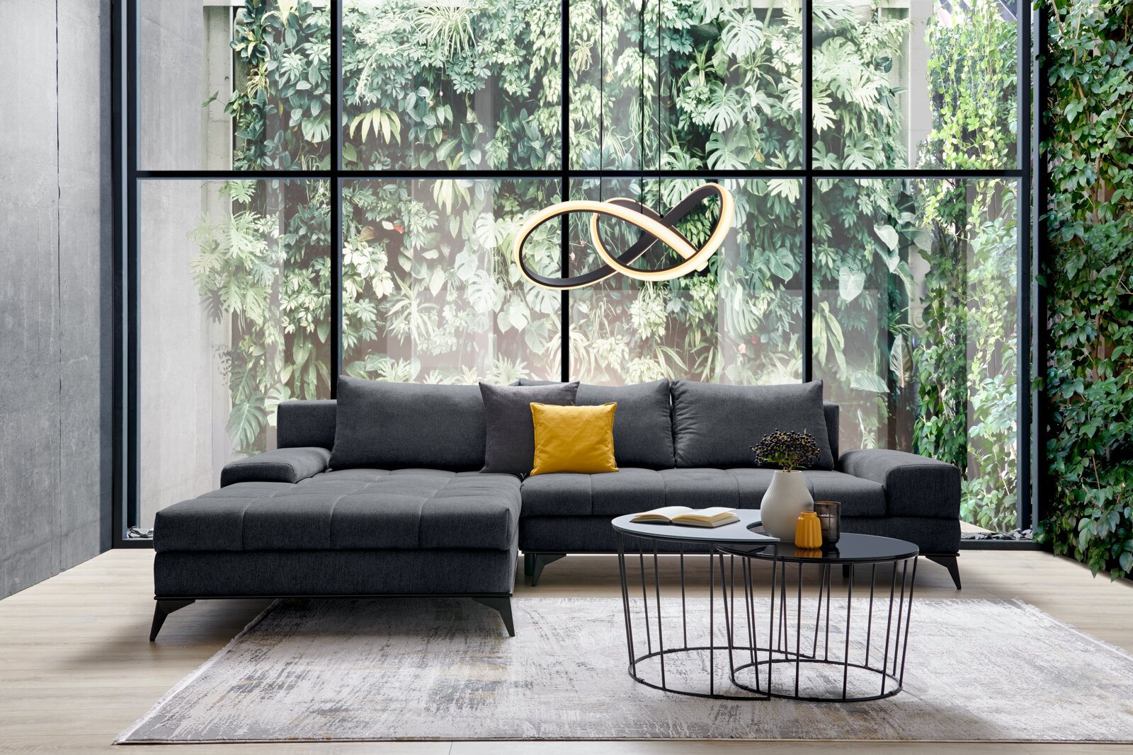 Graue Rundecke Ottli in modernem Wohnzimmer mit großen Fenstern und Pflanzen im Hintergrund, aus frontaler Perspektive fotografiert.