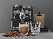 Ein Glas mit Cappuccino und ein leeres Glas auf einem Tisch, umgeben von Kaffeebohnen, mit einer Espressomaschine im Hintergrund. Perspektive von vorne.