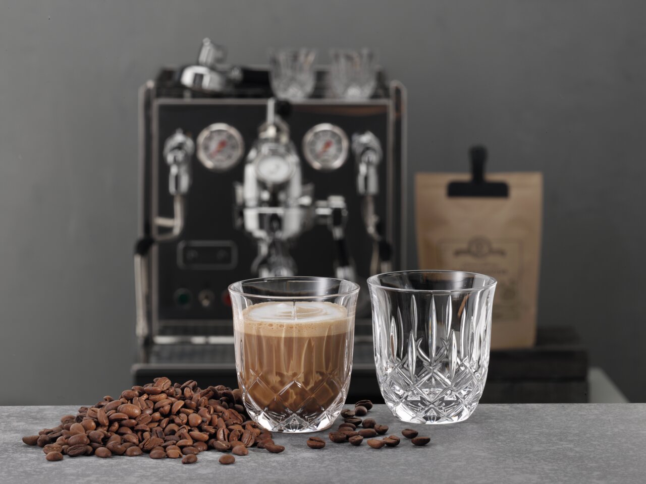 Ein Glas mit Cappuccino und ein leeres Glas auf einem Tisch, umgeben von Kaffeebohnen, mit einer Espressomaschine im Hintergrund. Perspektive von vorne.
