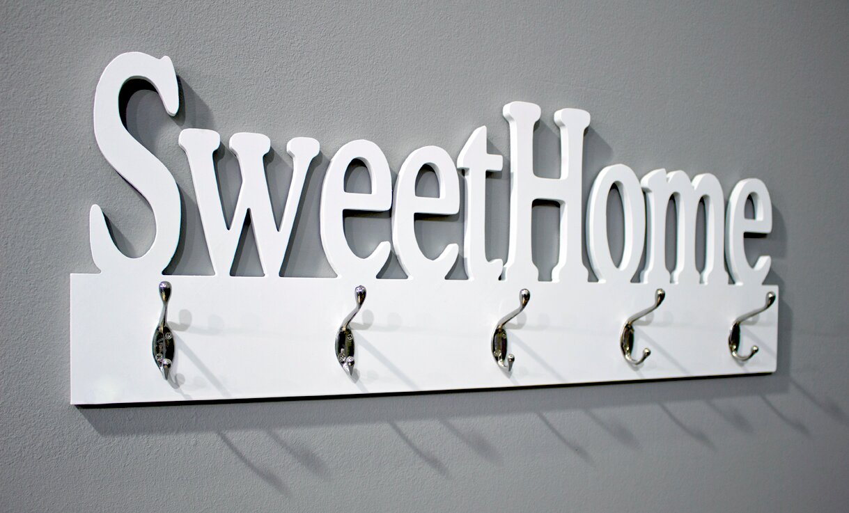 Weiße Wandgarderobe mit Schriftzug „Sweet Home“ und fünf silbernen Haken an grauer Wand, schräg von vorn links fotografiert.