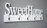 Weiße Wandgarderobe mit Schriftzug „Sweet Home“ und fünf silbernen Haken an grauer Wand, schräg von vorn links fotografiert.