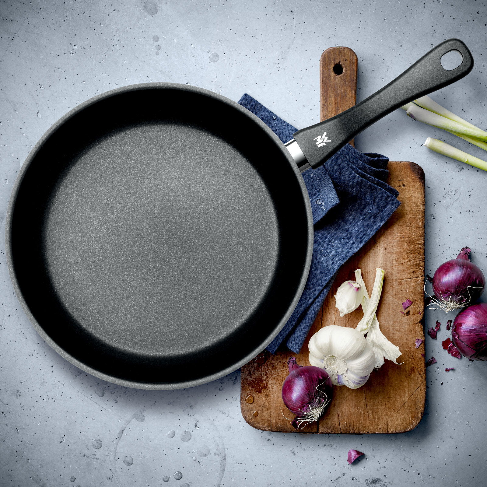 Stielpfanne 28cm aus der Vogelperspektive auf einem Schneidebrett mit Knoblauch und roten Zwiebeln