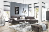 Ecksofa in einem modernen Wohnzimmer mit grauem Stoffbezug, dekoriert mit Kissen, im Vordergrund ein Couchtisch und ein Hocker, seitliche Perspektive.