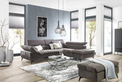 Ecksofa in einem modernen Wohnzimmer mit grauem Stoffbezug, dekoriert mit Kissen, im Vordergrund ein Couchtisch und ein Hocker, seitliche Perspektive.