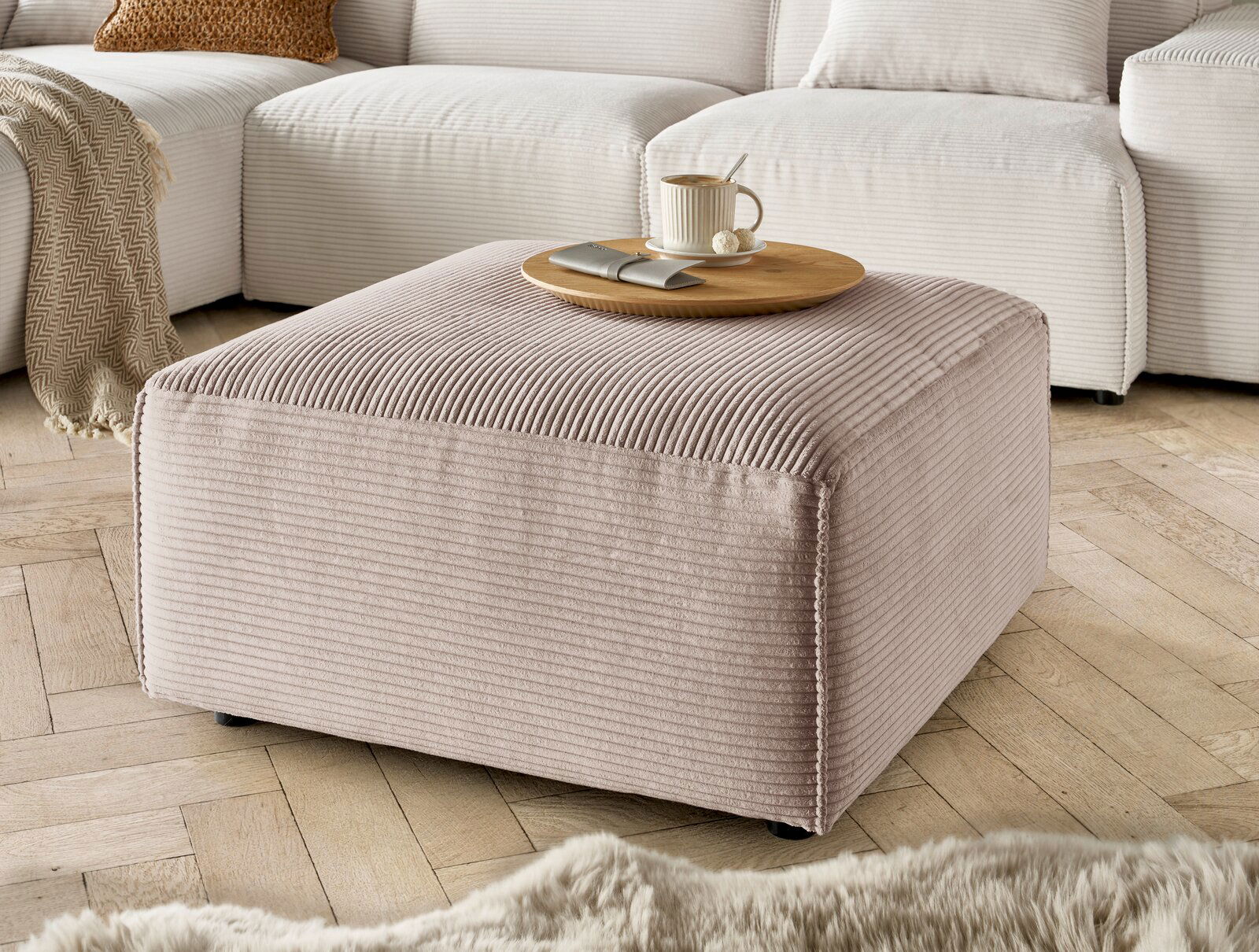 Hocker in Nahaufnahme, beige gestreifter Stoff, mit Tablett und Tasse darauf, auf Holzfußboden vor einem Sofa.