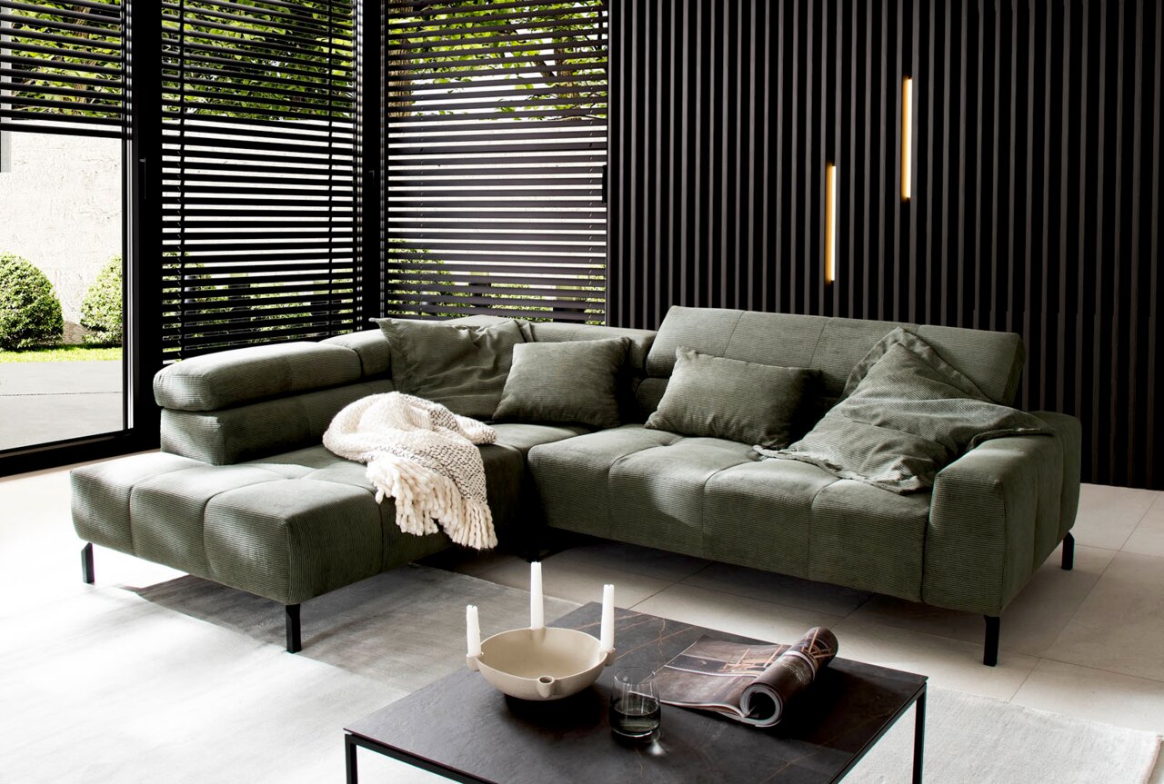 Lounge Handmade Ecksofa LH-CELSO Grüne Ecksofa in modernem Wohnzimmer mit großen Fenstern und stilvoller Dekoration, seitliche Perspektive.